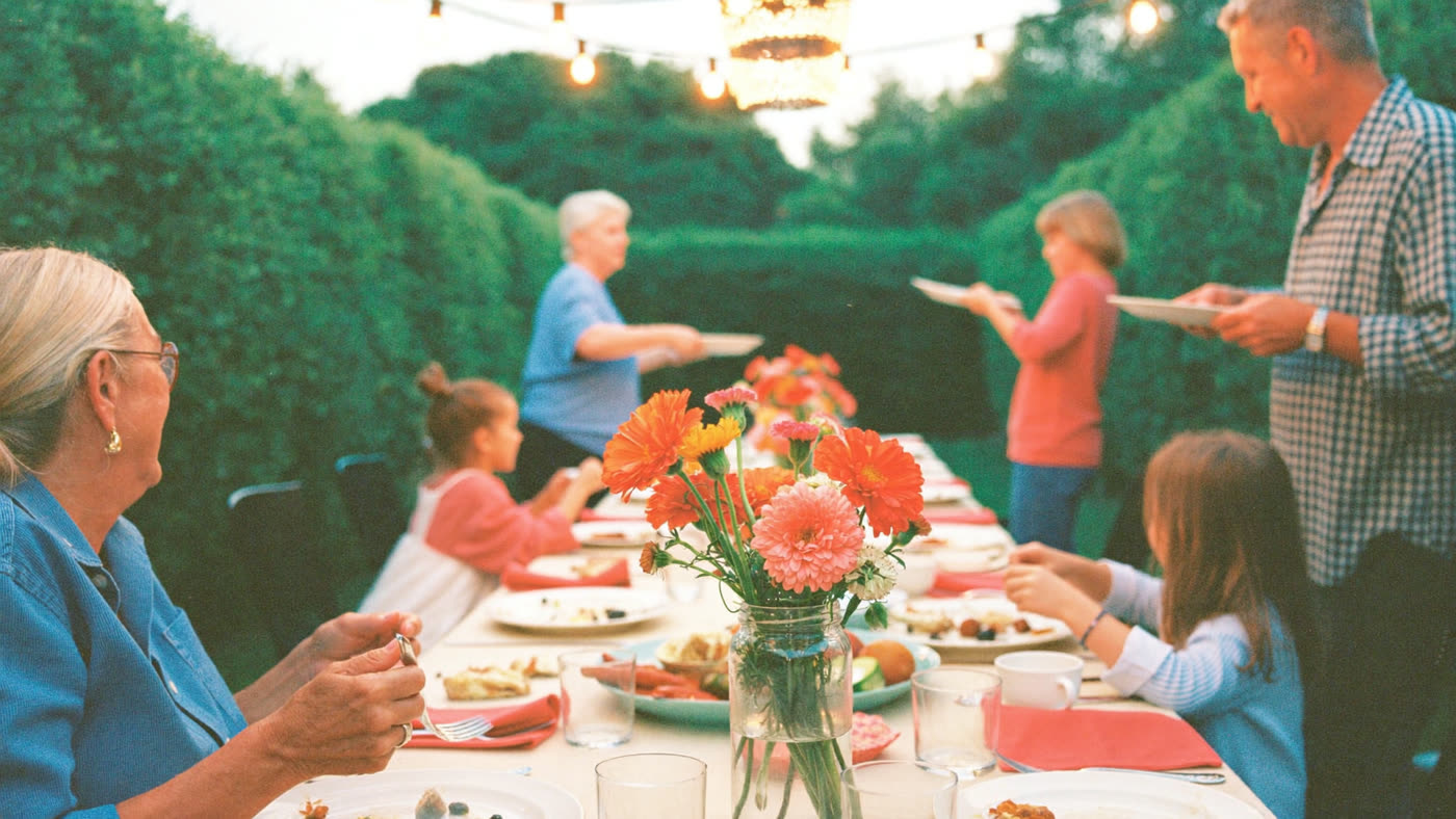 Three generations of a family sharing dinner at a long backyard table under string lights